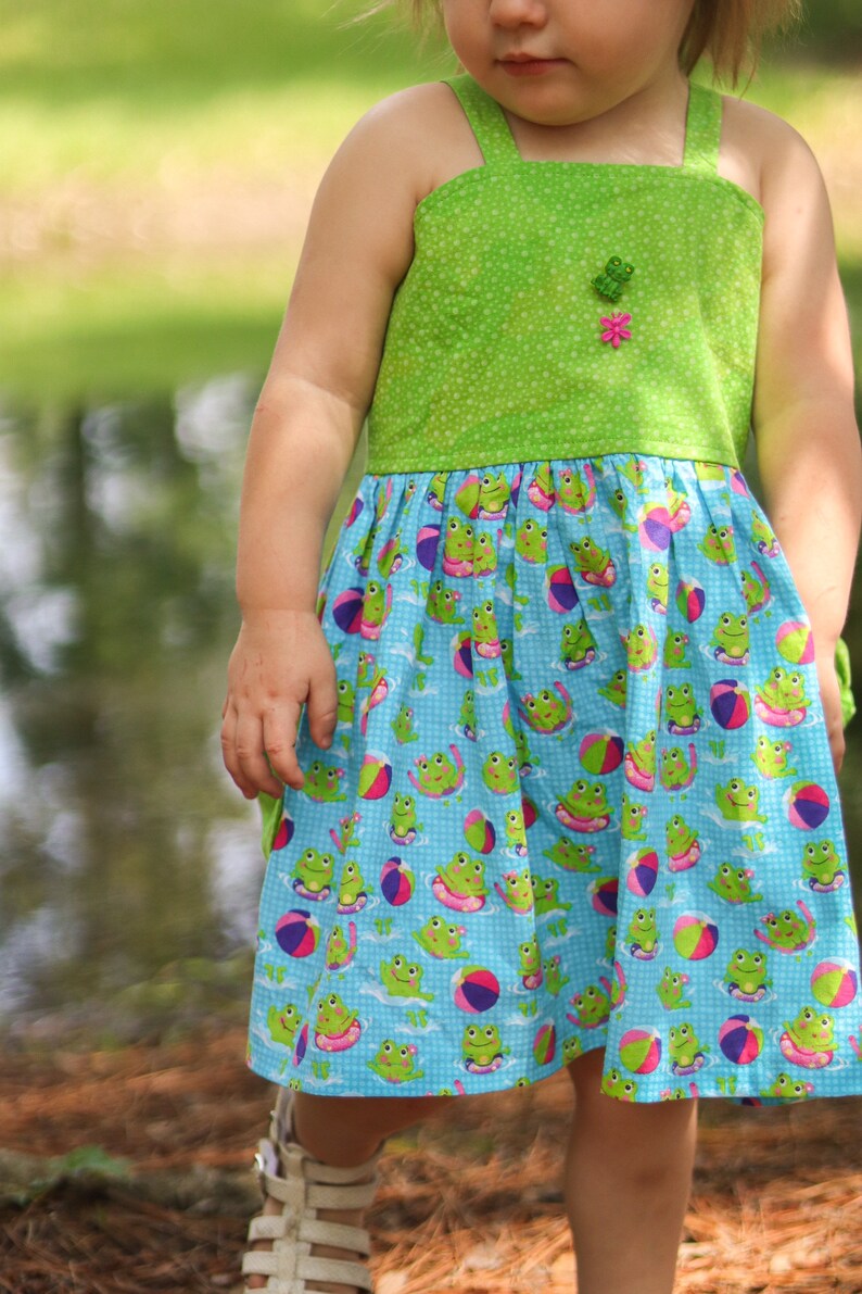 May include: A young girl wearing a green and blue sundress with a frog and beach ball pattern. The dress has a green bodice with a white polka dot pattern and a blue skirt with a frog and beach ball pattern.