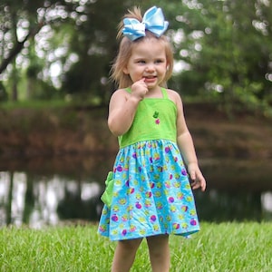 May include: A young girl wearing a green and blue dress with a frog and beach ball pattern. She is wearing a light blue bow in her hair and is standing in a grassy field.