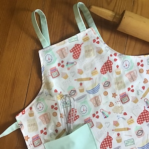 May include: A white apron with a light blue trim and a pocket. The apron has a pattern of baking-themed illustrations, including cupcakes, measuring cups, rolling pins, and hearts.