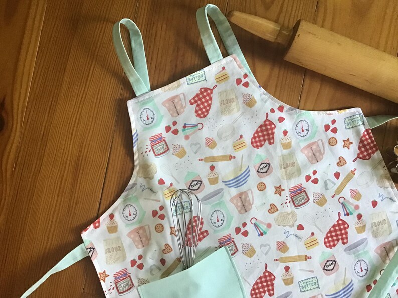 May include: A white apron with a light blue trim and a pocket. The apron has a pattern of baking-themed illustrations, including cupcakes, measuring cups, rolling pins, and hearts.