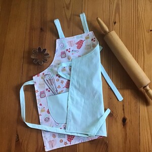 May include: A light blue children's apron with a pocket and a white background featuring a pattern of baking-themed illustrations. The apron is tied with two long ties and has a small pocket on the front. A wooden rolling pin is lying on a wooden surface next to the apron.