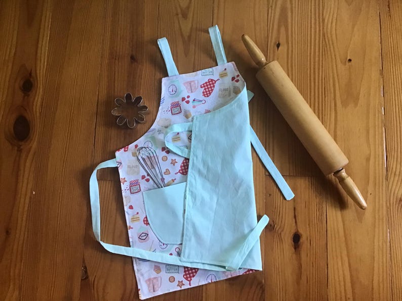 May include: A light blue children's apron with a pocket and a white background featuring a pattern of baking-themed illustrations. The apron is tied with two long ties and has a small pocket on the front. A wooden rolling pin is lying on a wooden surface next to the apron.