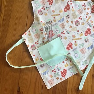 May include: A white apron with a light green pocket and a colorful pattern of baking-themed illustrations. The apron has adjustable straps and a tie closure.