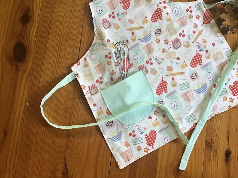 May include: A white apron with a light green pocket and a colorful pattern of baking-themed illustrations. The apron has adjustable straps and a tie closure.