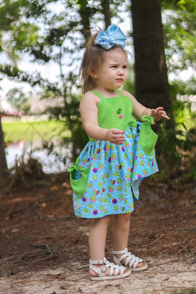 May include: A young girl wearing a green and blue dress with a frog and beach ball pattern. She is wearing white sandals and has a blue bow in her hair.