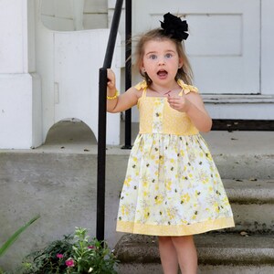 May include: A young girl wearing a yellow floral dress with a black bow in her hair stands on a set of concrete steps. She is looking at the camera with a surprised expression.