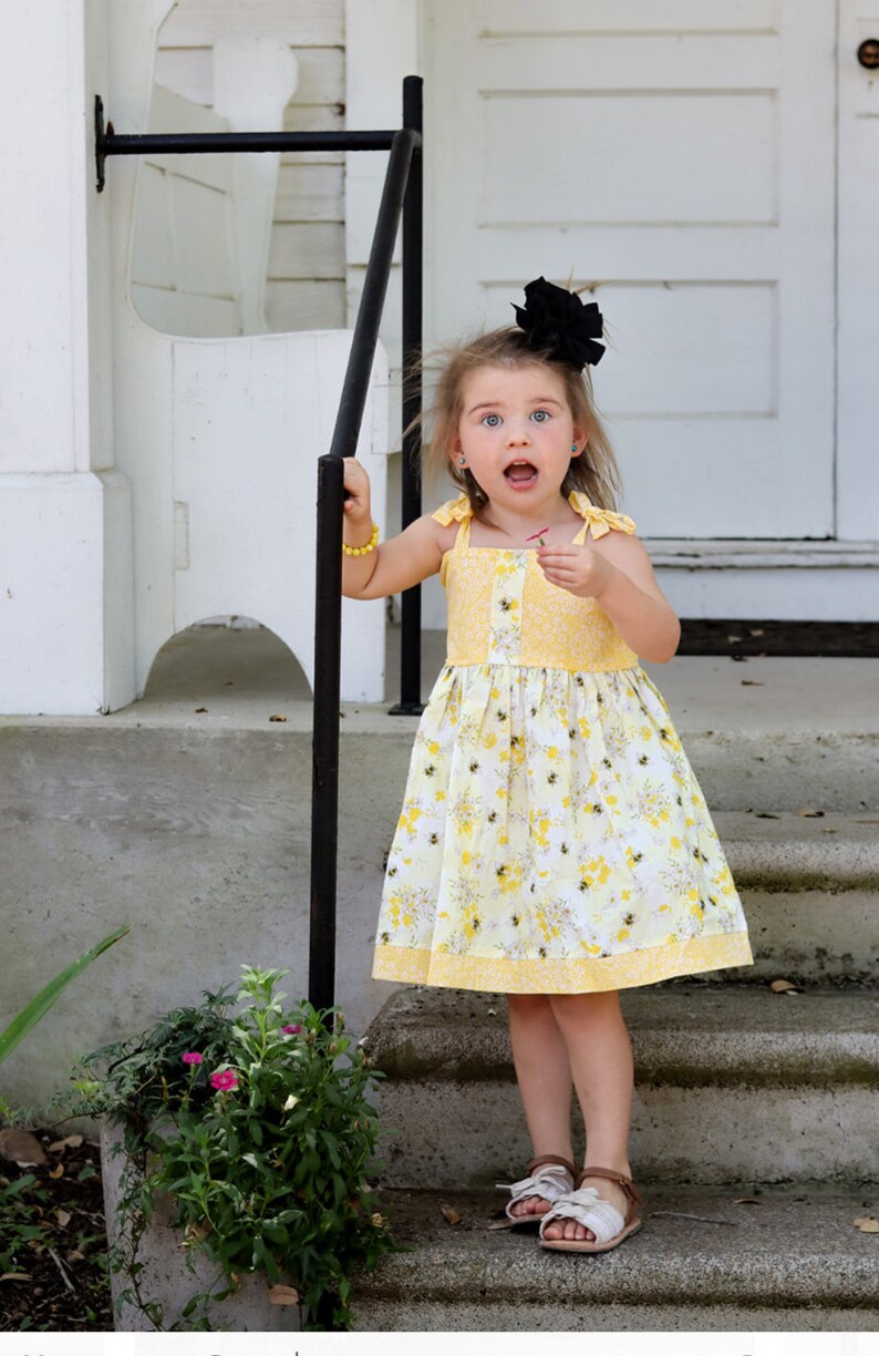 May include: A young girl wearing a yellow floral dress with a black bow in her hair stands on a set of concrete steps. She is looking at the camera with a surprised expression.