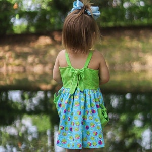May include: A young girl wearing a green and blue dress with a frog and beach ball pattern. The dress has a green bodice with a bow in the back and a blue skirt. The girl is standing in front of a blurred background of green trees.