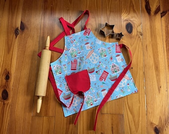 May include: A light blue apron with a festive gingerbread house pattern, red trim, and a red pocket. A wooden rolling pin and star-shaped cookie cutters are also visible, suggesting a baking theme.