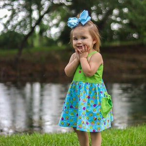 May include: A young girl wearing a blue and green dress with a frog print and a blue bow in her hair. She is standing in front of a pond and smiling.