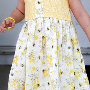 May include: A yellow and white sleeveless dress with a floral pattern and bees. The dress has a yellow trim and a yellow bow on each shoulder.