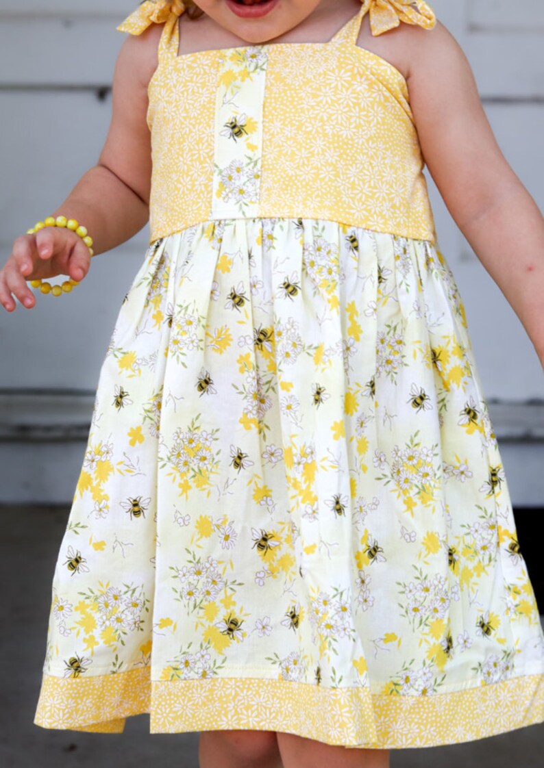 May include: A yellow and white sleeveless dress with a floral pattern and bees. The dress has a yellow trim and a yellow bow on each shoulder.