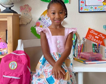 May include: A young girl wearing a pink dress with a white and blue patterned skirt, posing in front of a desk. A bright pink backpack with a white strap hangs on a chair. A sign reads "First Day of 3rd Grade."
