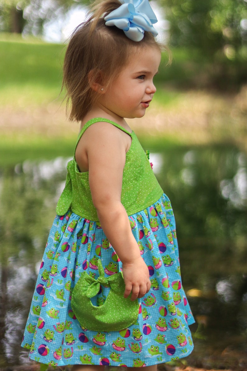 May include: A young girl wearing a green and blue dress with a frog and beach ball pattern. She is wearing a blue bow in her hair and holding a green fabric pouch.