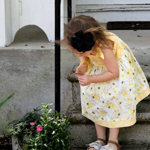 May include: A young girl wearing a yellow floral dress with a black bow in her hair is looking at a flower. She is standing on a set of concrete steps.
