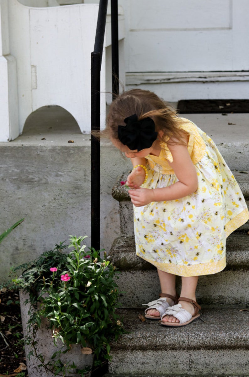 May include: A young girl wearing a yellow floral dress with a black bow in her hair is looking at a flower. She is standing on a set of concrete steps.