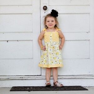 May include: A young girl wearing a yellow floral dress with a black bow in her hair stands in front of a white door. She is wearing brown sandals.
