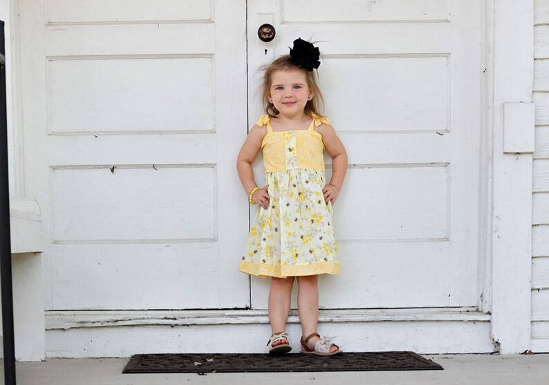 May include: A young girl wearing a yellow floral dress with a black bow in her hair stands in front of a white door. She is wearing brown sandals.