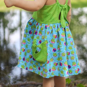 May include: A little girl wearing a green and blue dress with a frog and beach ball print. The dress has a green pocket and a bow in the back.