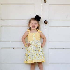 May include: A young girl wearing a yellow dress with a bee pattern and a black bow in her hair. She is standing in front of a white door with her hands on her hips.