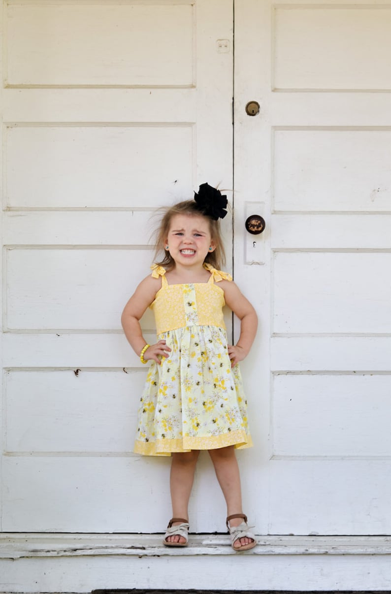 May include: A young girl wearing a yellow dress with a bee pattern and a black bow in her hair. She is standing in front of a white door with her hands on her hips.
