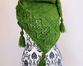 May include: A green knitted shawl with a lacy pattern and tassels. The shawl is draped over a white mannequin with a black base.