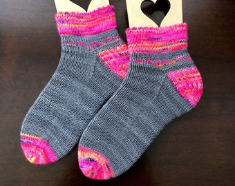 May include: A pair of gray and pink knit socks with a colorful stripe pattern at the top. The socks are displayed on wooden sock blockers with heart cutouts.