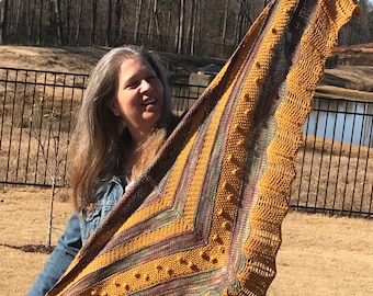 May include: A woman holds up a large, triangular shawl with a mustard yellow, green, and purple color scheme. The shawl has a textured pattern and a scalloped edge.