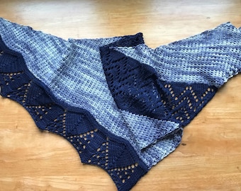 May include: A blue and white knitted shawl with a lacy border. The shawl is folded in half and laid flat on a wooden surface.