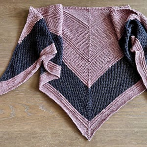 Könnte beinhalten: Ein gestrickter Schal in Dreiecksform mit einem farbblockierten Design. Der Schal hat einen dunkelgrauen Abschnitt und einen mauvefarbenen Abschnitt mit einem mauvefarbenen Rand. Die Textur des Strickwerks ist sichtbar. Der Schal wird auf einer Holzoberfläche präsentiert.