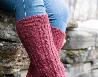 May include: A pair of burgundy knitted leg warmers with a cable knit pattern. The leg warmers are worn over blue jeans.