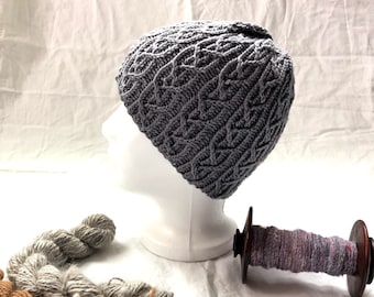 May include: A gray knitted hat with a textured pattern, displayed on a white mannequin head. Three skeins of yarn are visible in the foreground, one brown, one gray, and one light gray.