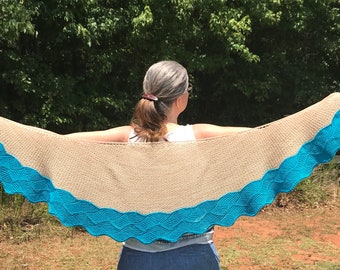 May include: A beige knitted shawl with a teal blue scalloped border. The shawl is draped over the shoulders of a person standing in a grassy field.
