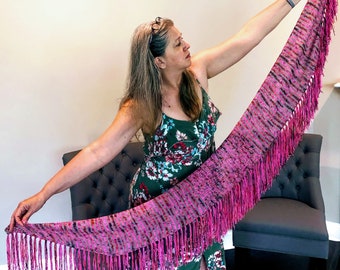 May include: A woman wearing a green floral dress holds a pink and purple fringed shawl with a textured pattern.