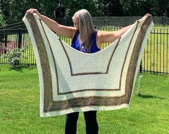 May include: A white knitted shawl with a green and purple border. The shawl is held up by a person wearing a blue tank top and black pants.