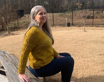 May include: A woman wearing a yellow knitted sweater with a cable knit design on the sleeves, sitting on a wooden bench outdoors.