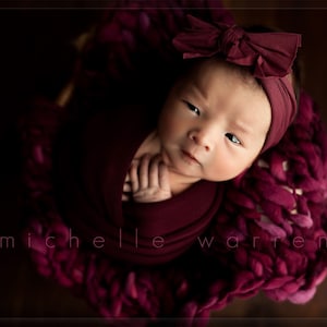 May include: A newborn baby girl is swaddled in a burgundy knitted blanket and wearing a burgundy headband with a matching bow. The baby is looking up at the camera with a serious expression.