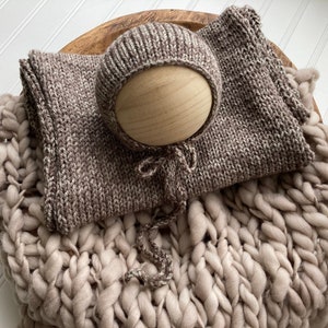May include: A brown and beige knitted baby hat and wrap set on a cream chunky knit blanket. The hat has a small opening at the top for the baby's head.