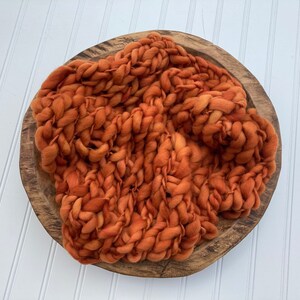 May include: A chunky knit blanket in a burnt orange color, displayed in a wooden bowl. The blanket is made of thick, soft yarn and has a textured, woven appearance.