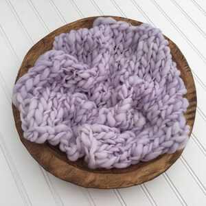 May include: A light purple knitted baby prop in a wooden bowl. The knitted prop is made of chunky yarn and has a soft, fluffy texture.