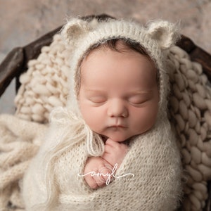 May include: A newborn baby is sleeping soundly in a white knitted hat with bear ears and a white knitted blanket. The baby is lying in a brown wooden basket.