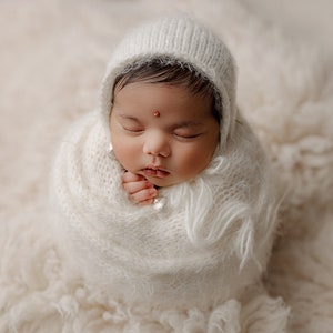 May include: A newborn baby is swaddled in a white knitted blanket and wearing a white knitted hat. The baby has a small red dot on their forehead.