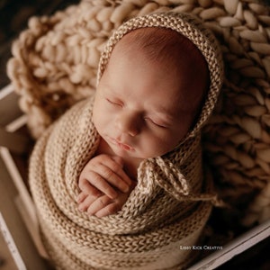 May include: A newborn baby is swaddled in a beige knitted blanket and a matching knitted hat. The baby is sleeping peacefully with their hands clasped together. The baby is lying in a white wooden box.