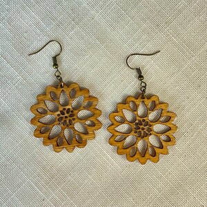May include: A pair of wooden earrings with a delicate, intricate floral design. The earrings are a light brown color and have a simple hook closure.
