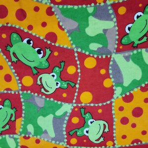 May include: A colorful fabric featuring a patchwork design with green frogs, red squares, yellow circles, and green camouflage shapes. The frogs have big eyes and are smiling. The fabric is likely for children's items.