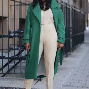 Pu&ograve; includere: Una persona indossa un lungo cappotto verde smeraldo sopra una camicetta e pantaloni color crema. I pantaloni sono risvoltati alle caviglie e indossa sandali neri con tacco alto. Indossa anche occhiali da sole.
