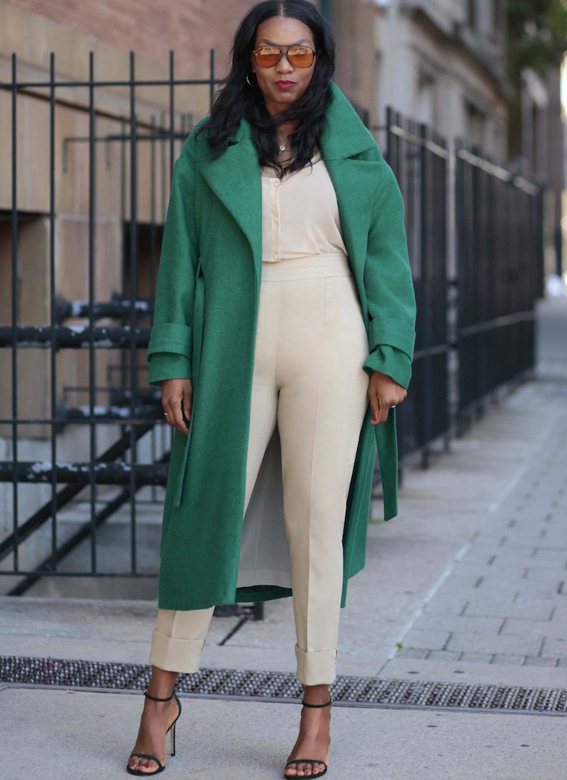 Pu&ograve; includere: Una persona indossa un lungo cappotto verde smeraldo sopra una camicetta e pantaloni color crema. I pantaloni sono risvoltati alle caviglie e indossa sandali neri con tacco alto. Indossa anche occhiali da sole.
