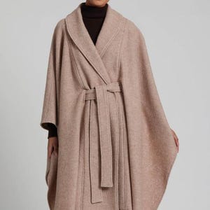 May include: A light brown cape with a shawl collar and a matching belt. The cape is made of a soft, textured fabric and has a wrap-around design. The model is wearing a dark brown turtleneck underneath.