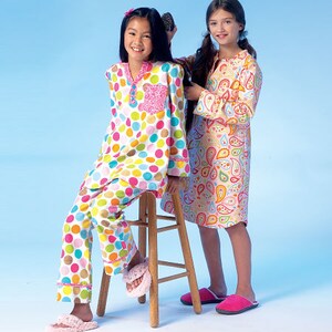 May include: Two girls in colorful pajamas. One girl is seated on a wooden stool wearing a pajama set with large polka dots in various colors. The other girl is standing, wearing a paisley-patterned nightgown. Both girls are wearing slippers.
