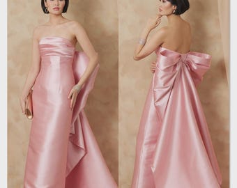 Può includere: Busta per cartamodello Vogue Patterns V2177 che mostra un abito lungo senza spalline in raso rosa. L'abito presenta un corpetto aderente con pieghe orizzontali e un grande fiocco decorativo sul retro che si estende in un lungo strascico.